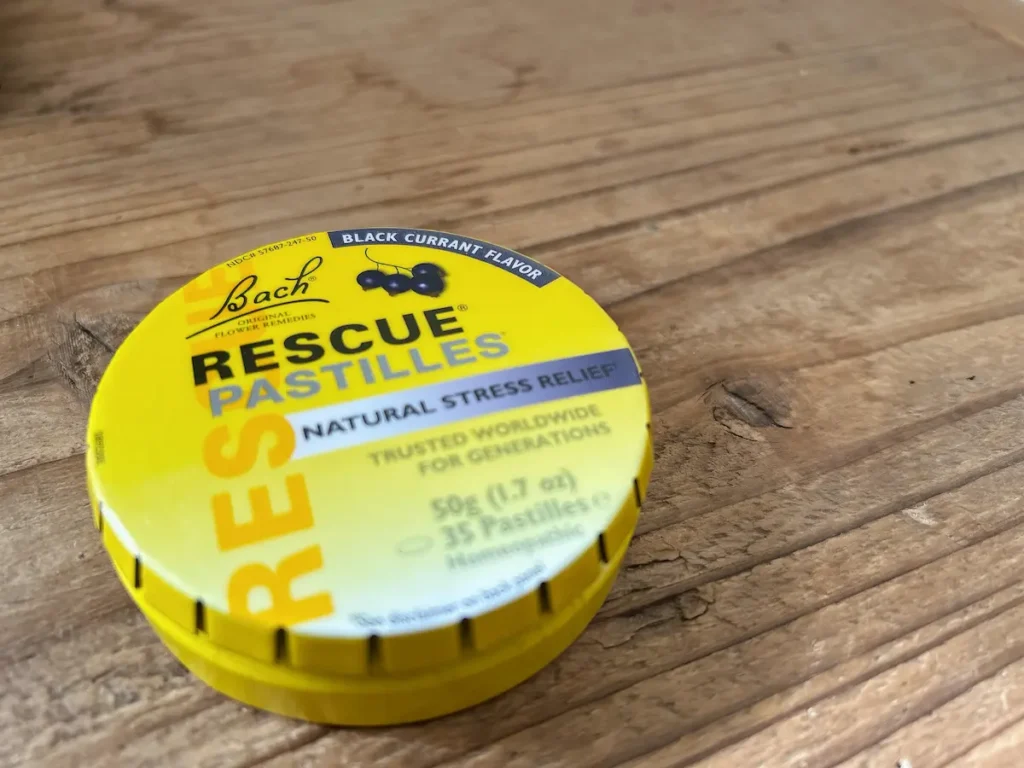 木目調のテーブルの上に置かれた、BACHレスキューレメディ・パスティル（ブラックカラント味）の丸い黄色い缶。蓋には黒スグリのイラストと「RESCUE PASTILLES」の文字がある。