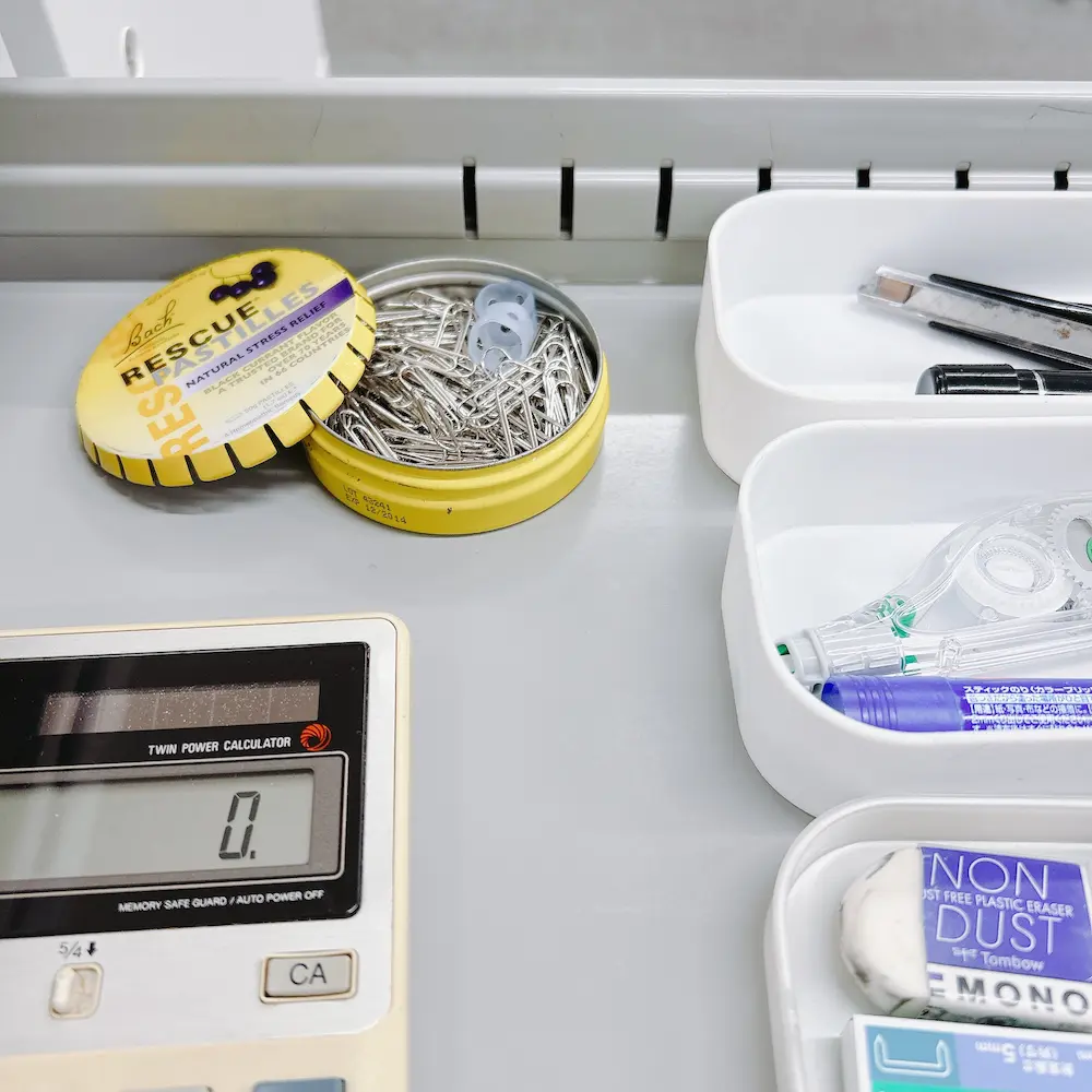 グレーの職場のデスクの引き出しが開いており、その中にBACHレスキューレメディの空き缶（黄色）が置かれている。缶の中には銀色のクリップと、小さな透明なプラスチックの小物が数個入っている。