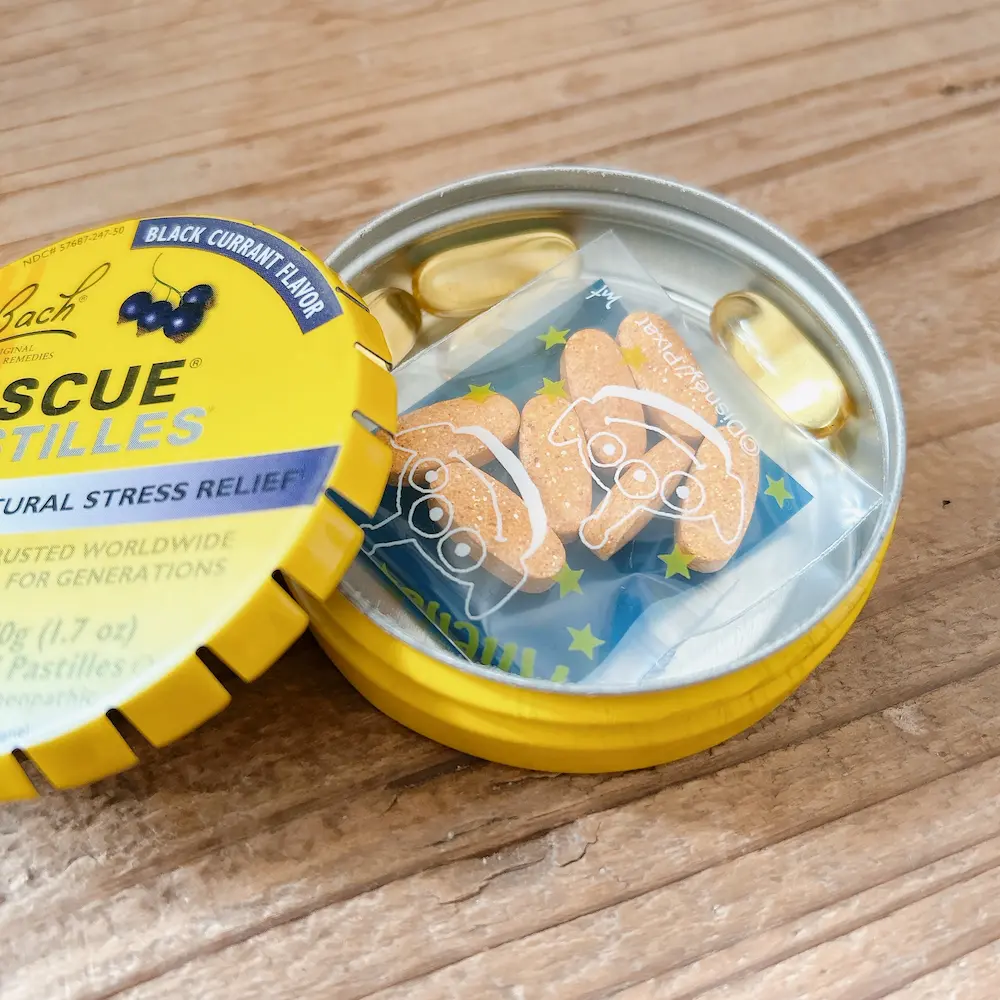 木目調のテーブルの上にあるBACHレスキューレメディの缶の蓋が少し開いており、中が見える。缶の中には、個包装されたオレンジ色のタブレット（Disney/Pixarのキャラクターが描かれている）と、ゴールドのカプセルが数粒入っている。